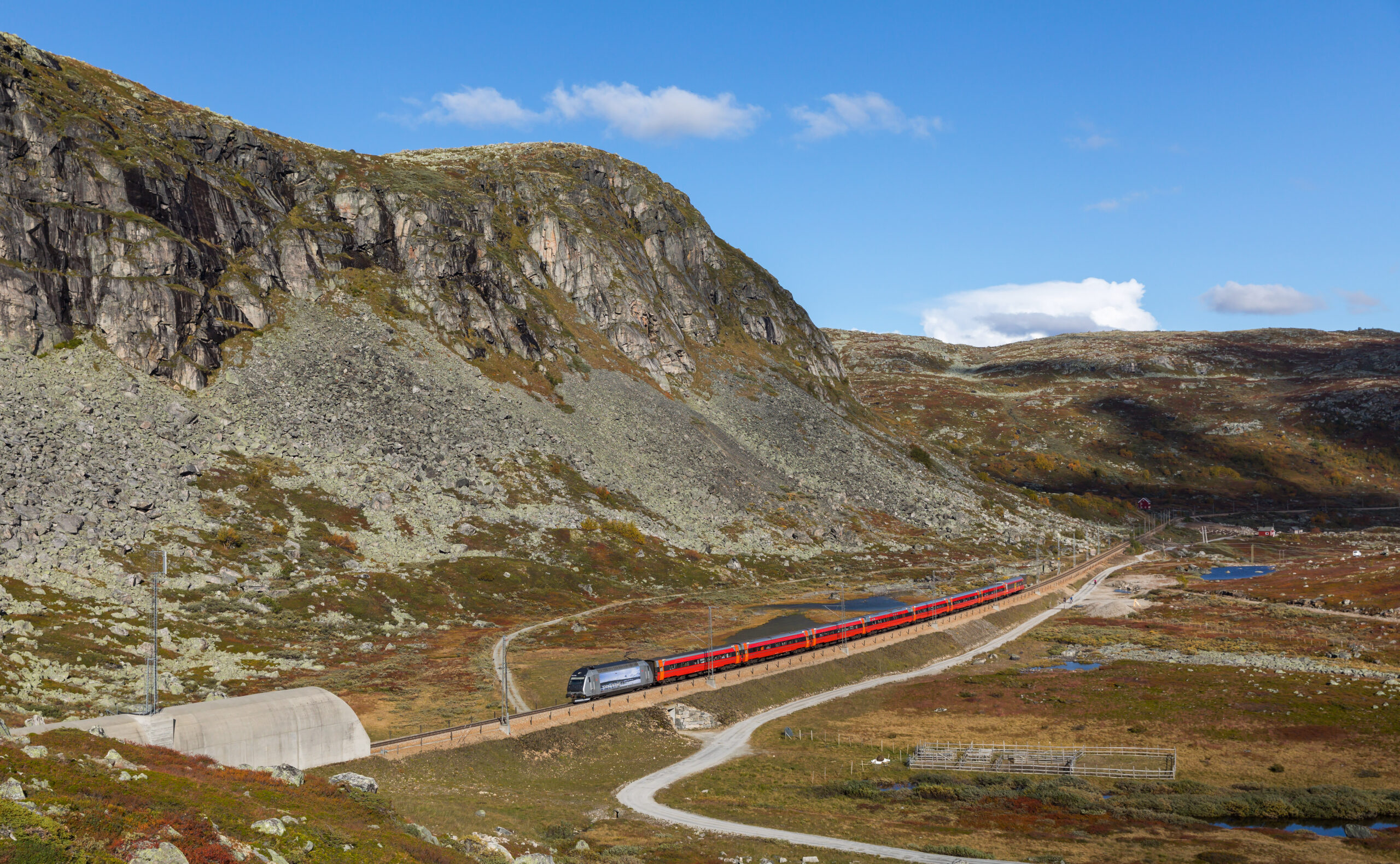 E-Lok mit roten Reisezugwagen auf der Bergenbahn in der felsigen Hardangervidda bei Haugastøl, Norwegen